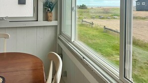 Ein Zimmer mit großem Fenster, einem Holztisch, einem weißen Stuhl und einer kleinen Pflanze auf einem Regal.