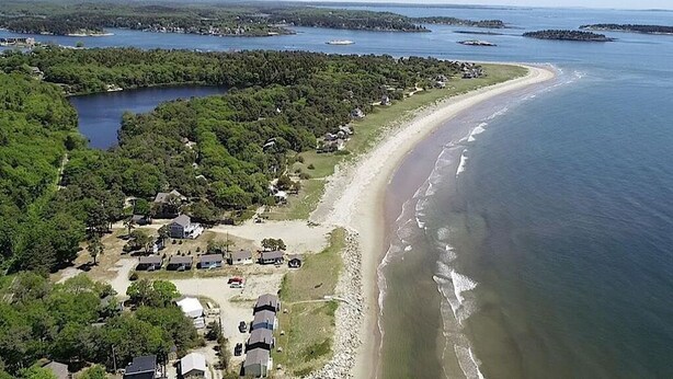 Eine Vogelperspektive zeigt eine Küstenlandschaft mit Strand, einer kleinen Siedlung mit Häusern und Fahrzeugen sowie mehreren Wasserflächen.