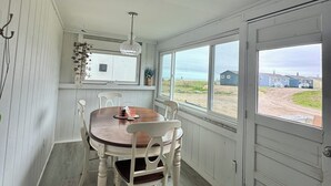 Ein Essbereich mit einem Holztisch, weißen Stühlen, einer Hängeleuchte und Blick auf den Strand durch große Fenster.