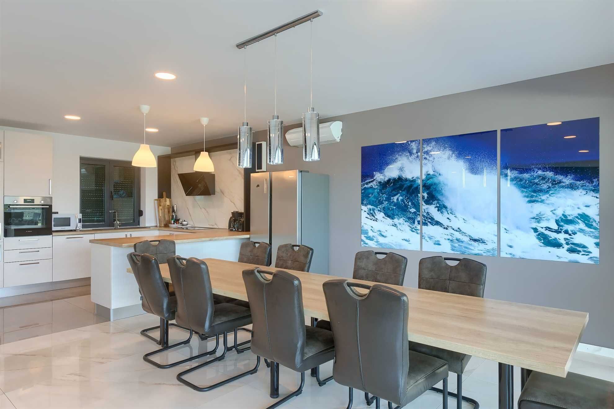 Een moderne keuken met een centraal eiland, een eethoek met een lange tafel en stoelen, en een groot schilderij van een oceaan golf aan de muur.