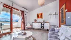 A modern living room with a sofa, a coffee table, and a dining area with a cabinet. There is a view of mountains through the glass door. - Lori Red - Cozy Hideaway for Two with sea view 🌊 (Baška)