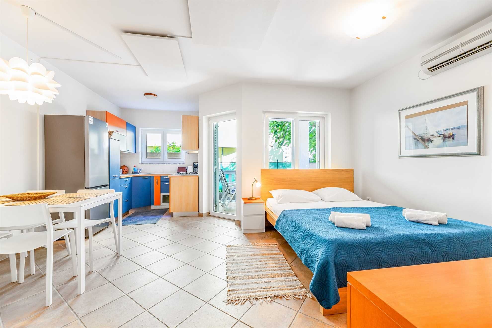 A compact hotel room with a bed, a desk, a kitchenette, and a view of greenery.