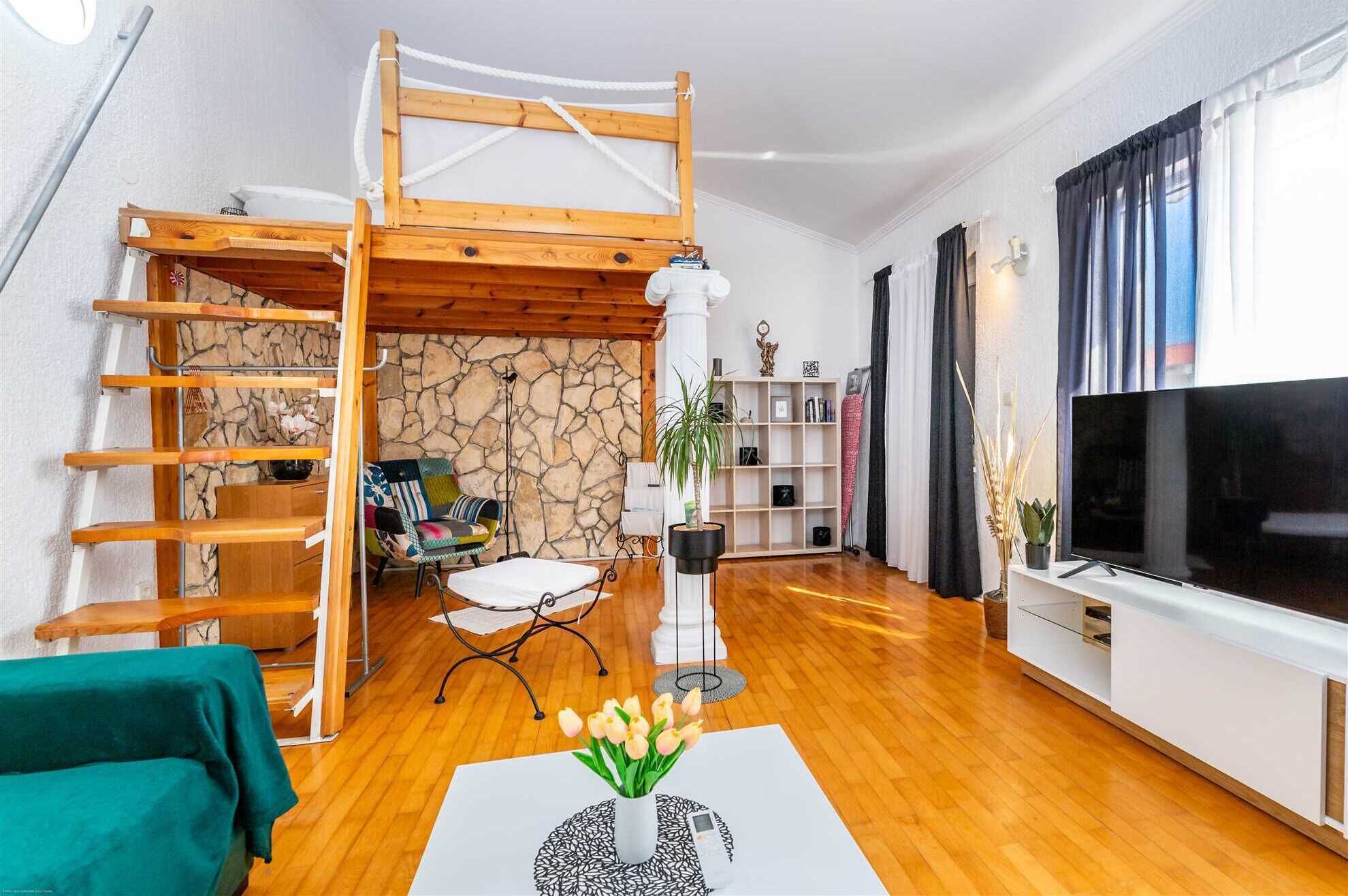 A cozy living room with a wooden loft bed, a TV, a bookshelf, and a stone wall.