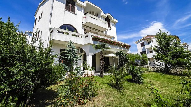 A two-story building with a white exterior, multiple balconies, and a garden with various plants and flowers. - Apartment Bozo Baska on the ground floor with terrace (Baška)