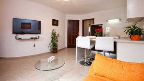 A modern living room with a flat-screen TV, a glass coffee table, a white sofa, and a dining area with a white chair and a small table. - Apartment Bozo Baska on the ground floor with terrace (Baška)