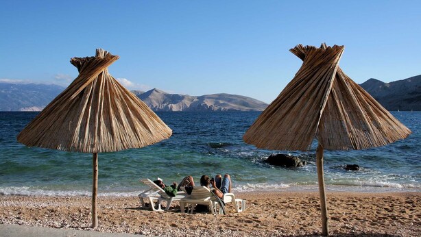 Een strand met twee rieten parasols, een kiezelstrand en een bergzicht op de achtergrond.