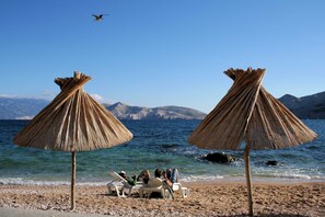 Ein Strand mit zwei Strohdächern, Kiesstrand und Blick auf eine Berglandschaft im Hintergrund.
