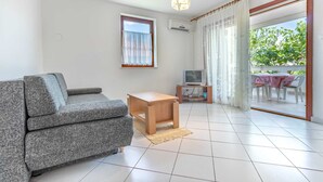 A living room with a grey sofa, a wooden coffee table, a television on a stand, and a sliding glass door leading to an outdoor area with a table and chairs. - Family apartment Antica A4 (Baška)