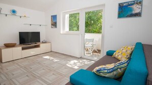 Ett modernt vardagsrum med en platt-TV, en soffa med mönstrade kuddar, en matplats med stolar och en balkong med utsikt över grönområden.