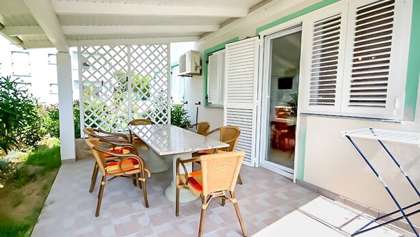 Een overdekt terras met een tafel en stoelen, een witte latten schutting en een schuifdeur naar een keuken.