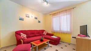 Een woonkamer met een rode bank, een houten salontafel, een televisie op een standaard en een raam met dunne gordijnen.