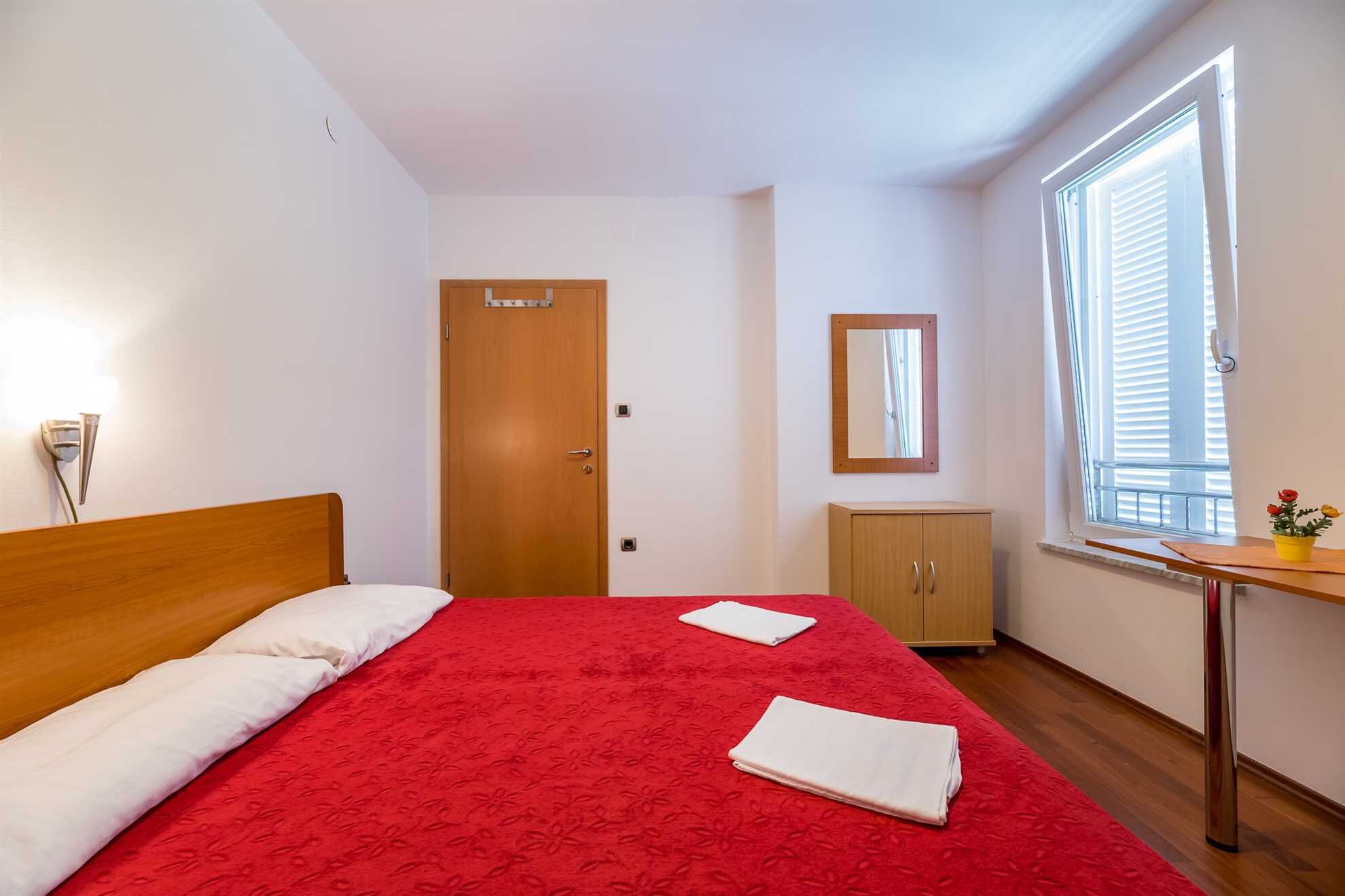 Une chambre avec un lit en bois, une couvre-lit rouge, une petite commode en bois, une table de chevet avec un miroir et une fenêtre avec des stores.