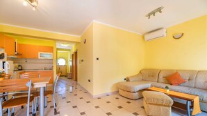 A living room with a sofa, ottoman, and a coffee table. There is a dining area with a table and chairs. The room has yellow walls and a tiled floor. - Apartment Nada 2 with air condition and wifi internet (Baška)