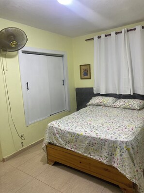Une chambre à coucher avec un lit, un ventilateur de plafond, une fenêtre avec des rideaux et un tableau accroché au mur.