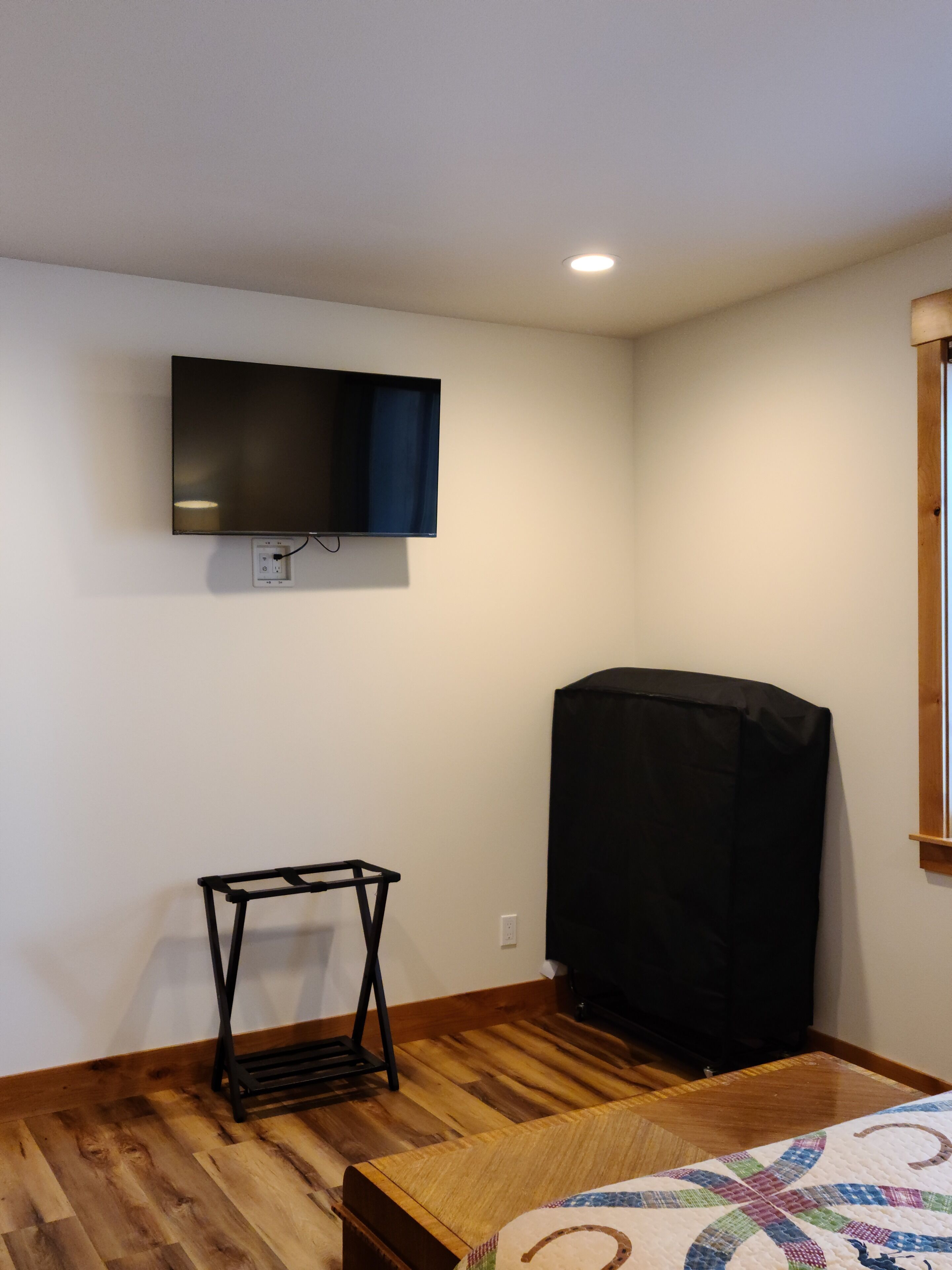 A room with a flat-screen TV mounted on the wall, a wooden floor, and a bed with a quilt.