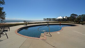 Una piscina con una escalera metálica, rodeada por una plataforma de concreto, con vista a la playa y al océano al fondo.