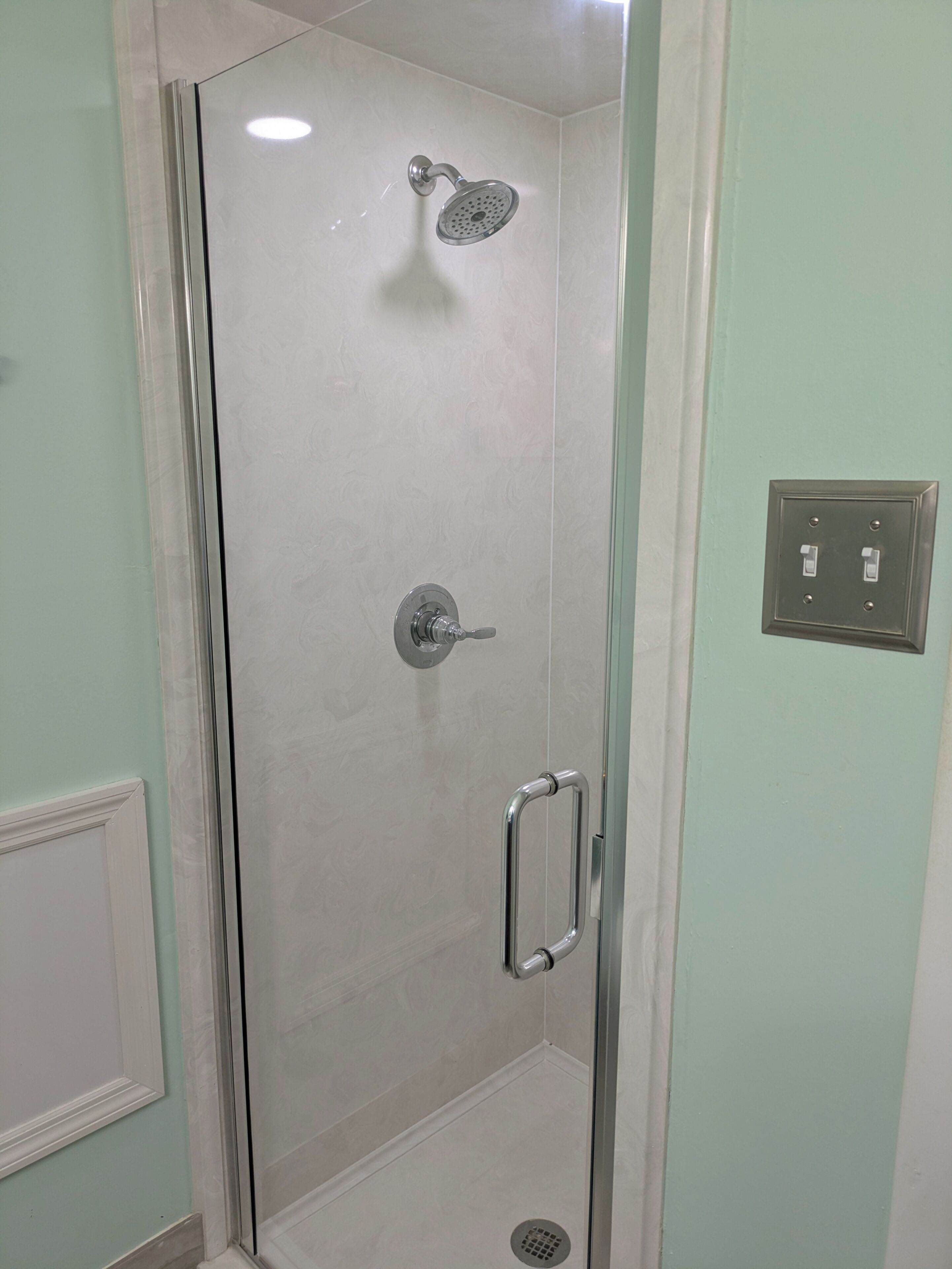 A glass-enclosed shower with a handheld showerhead, a wall-mounted control, and a handrail.