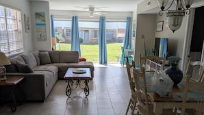 Una sala de estar con sofá, mesa de centro, mesa de comedor con sillas y vista al patio trasero.