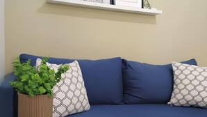 Un divano blu con cuscini decorati, un tavolino bianco tondo con una pianta e libri, e una parete con opere d'arte incorniciate.