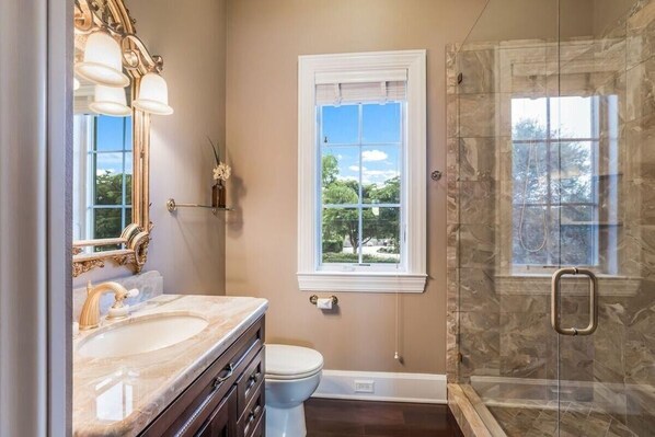 A bathroom with a marble sink, a toilet, a shower with a glass door, and a window with a view of trees.