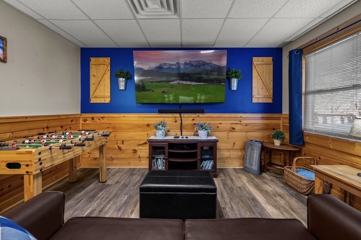 Quarto com mesa de pebolim, televisão de tela plana fixada numa parede azul, painéis de madeira e janela com persianas.