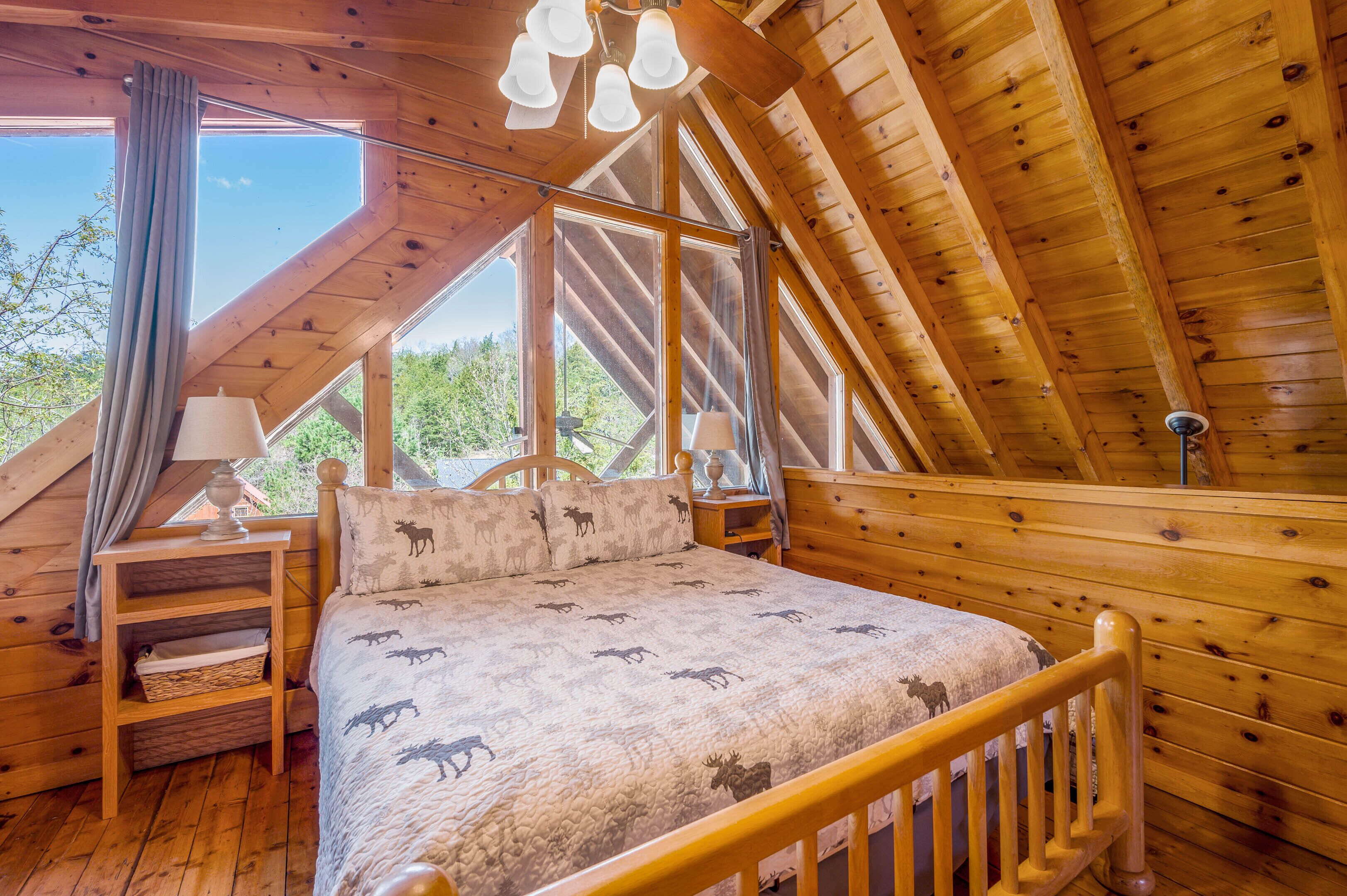 Une chambre à coucher en bois, avec un lit orné de motifs représentant des élans, une table de chevet, une lampe et une vue sur l’extérieur grâce à de grandes fenêtres.