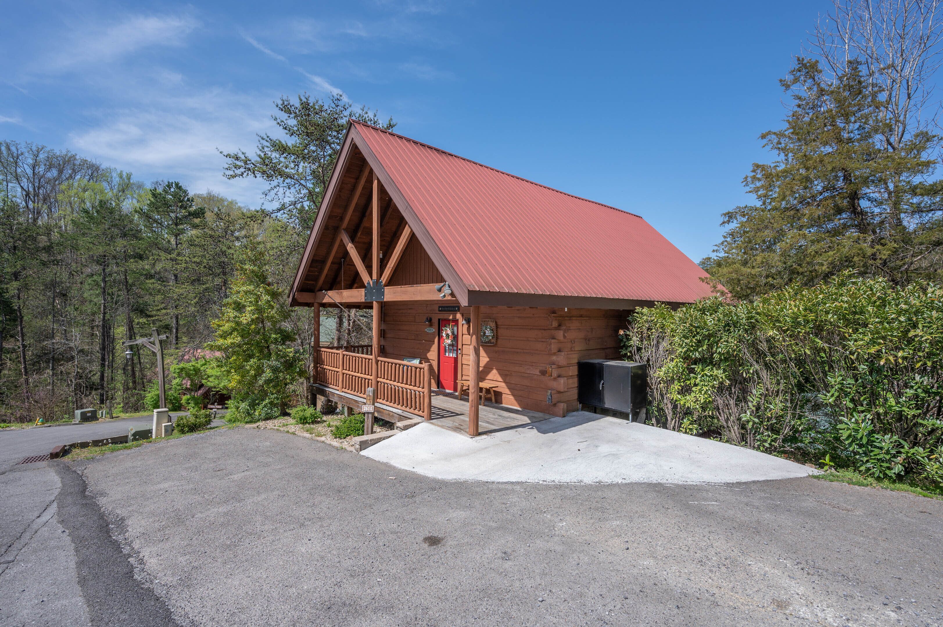A wooden cabin with a red roof, a porch, and a concrete driveway.