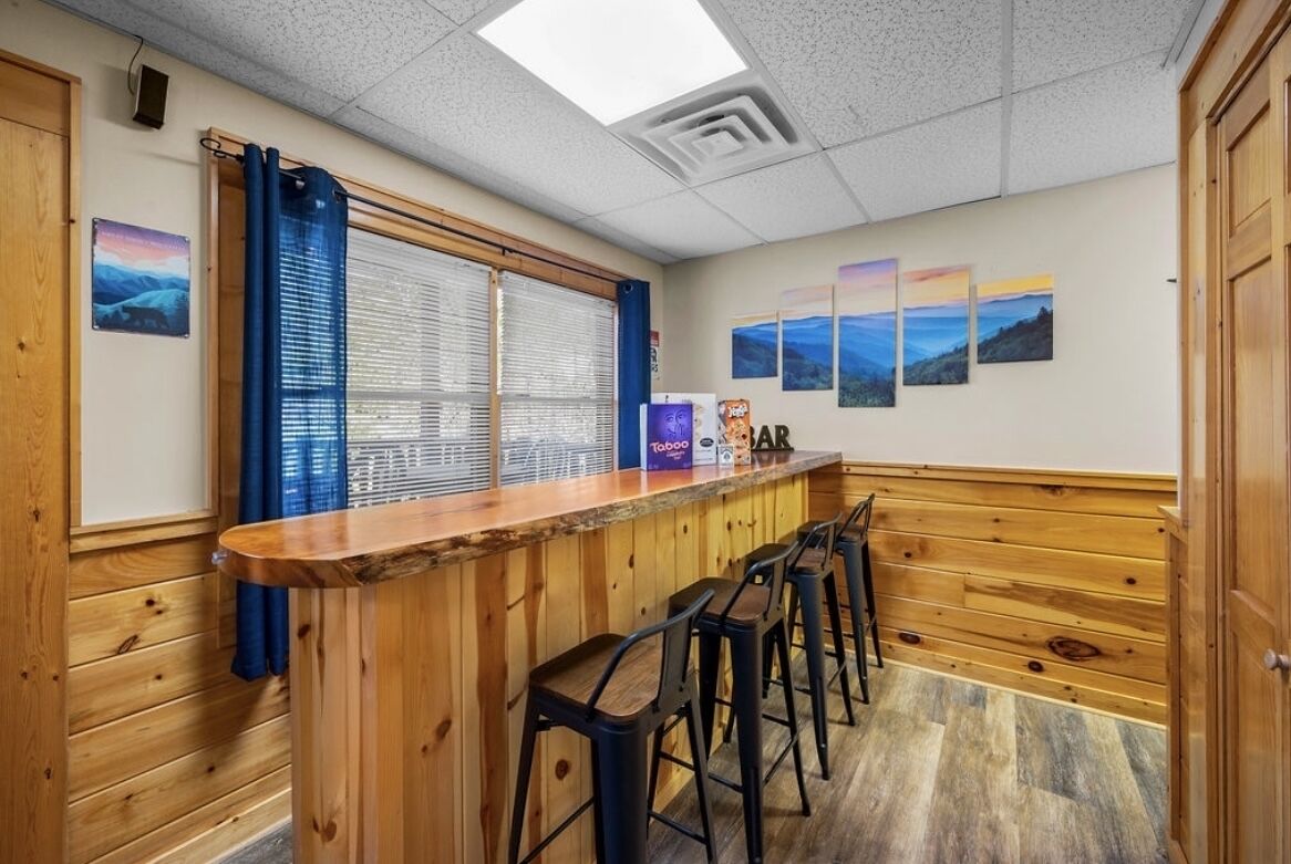 A bar area with wooden bar counter and stools, a window with blinds, and framed artwork on the wall.