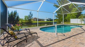 A covered outdoor swimming pool with lounge chairs, a table with a vase of flowers, and a view of a house and trees in the background. - Waterfront Cape Coral Villa w/ Heated Pool, 4 Bedrooms, Pet-Friendly (Cape Coral)