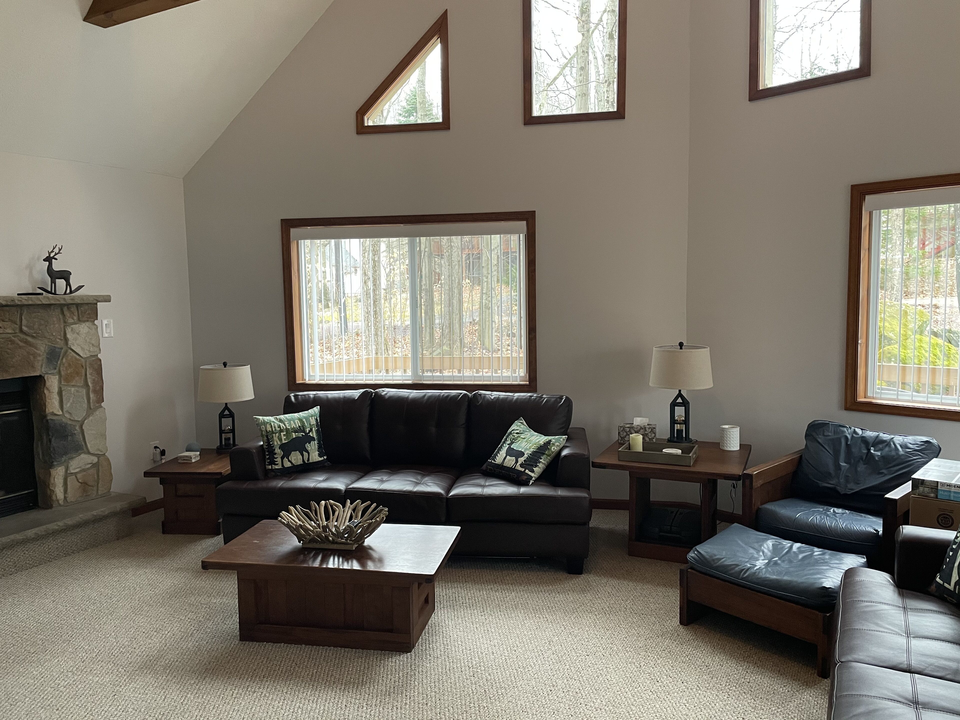 A living room with a stone fireplace, a leather sofa, a coffee table, and large windows with a view of trees.