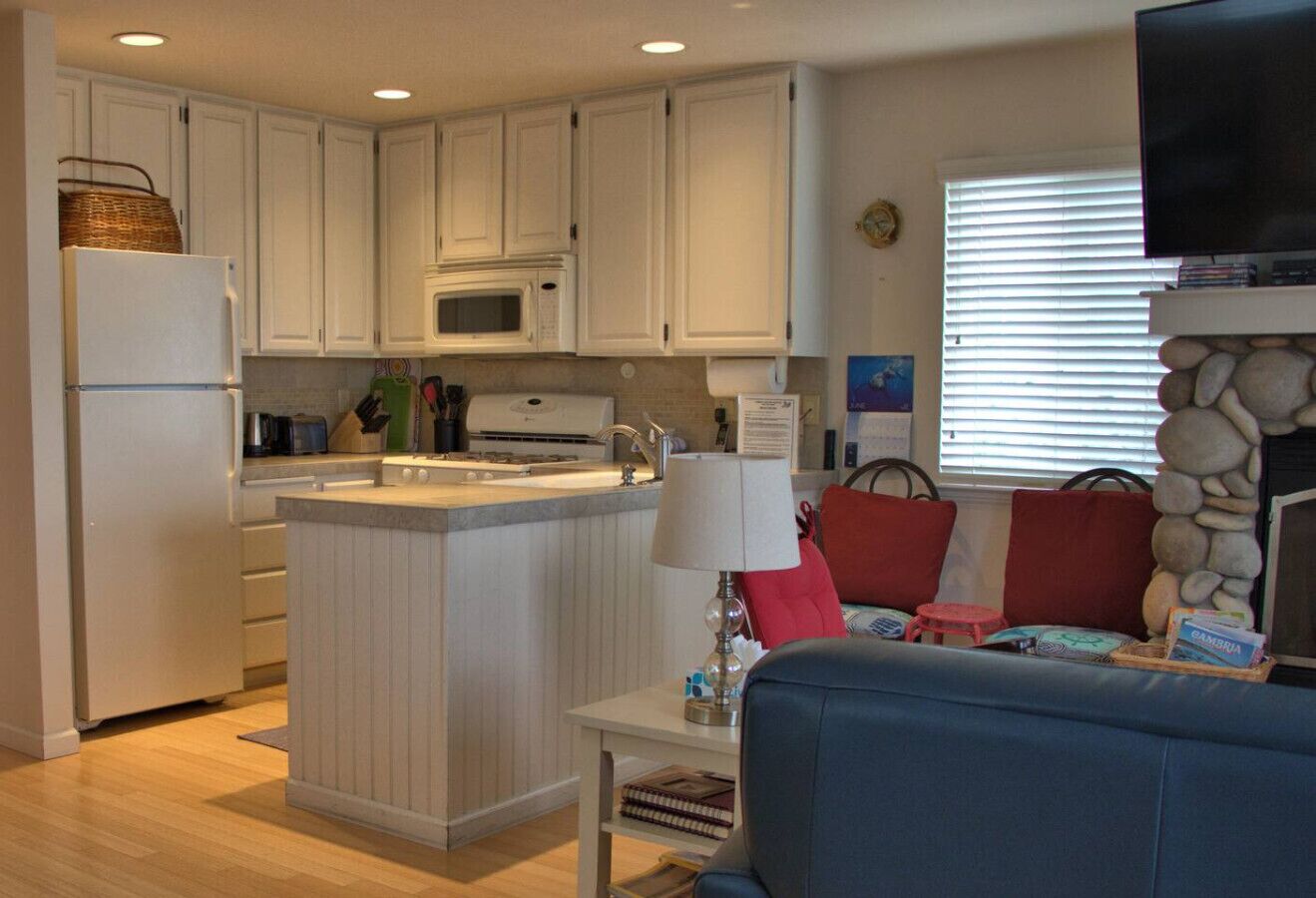 A compact kitchen with white cabinets, a microwave, and a refrigerator. There's a small island with a lamp and a red cushion. A stone wall and a television are visible in the background.