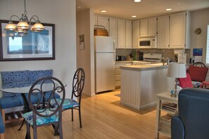 A compact kitchen with white cabinets, a central island, a dining area with a blue table and chairs, and a hanging light fixture.