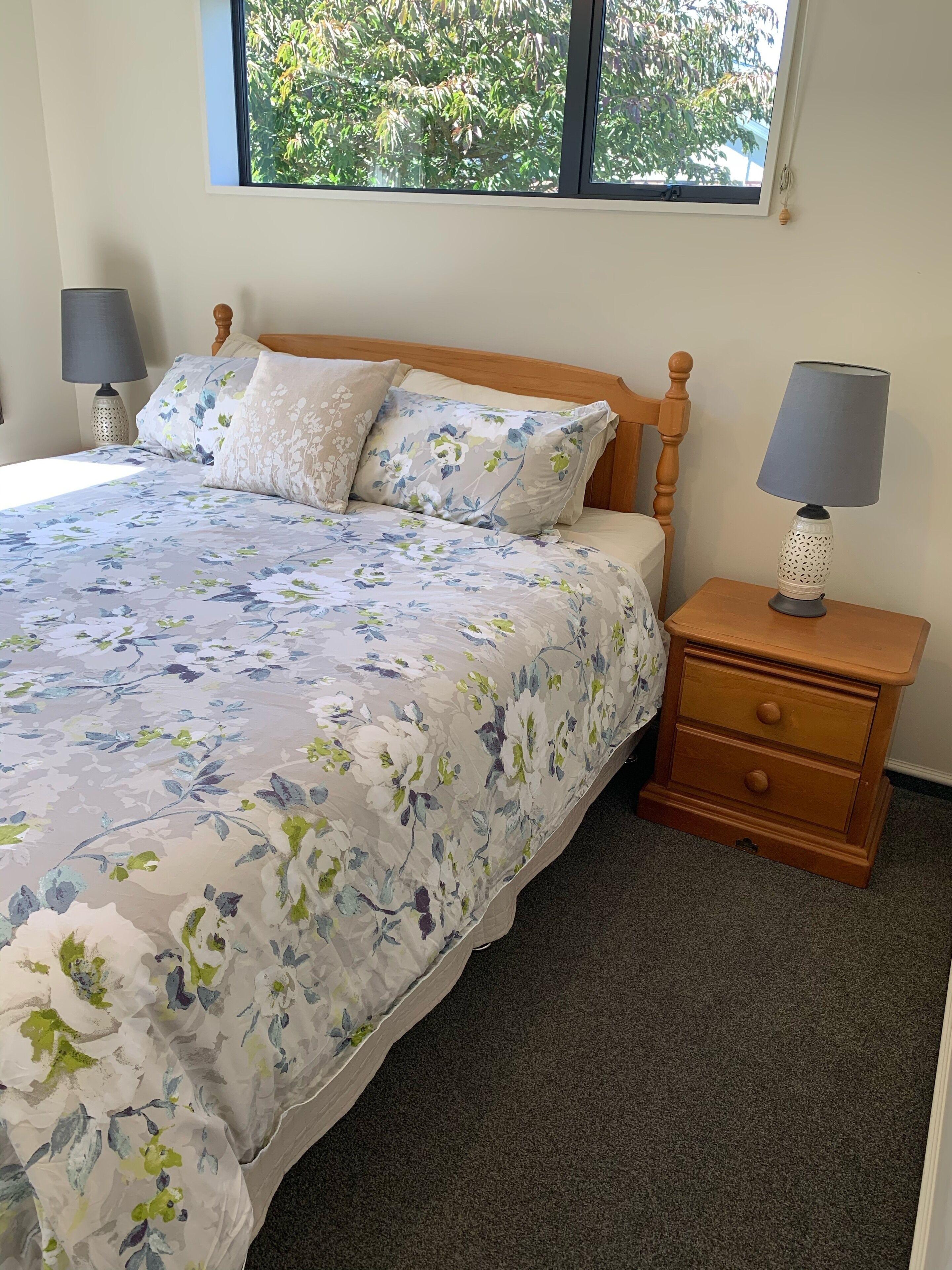 A bedroom with a floral bedspread, a wooden headboard, two bedside lamps, a nightstand with drawers, and a window with a view of trees.