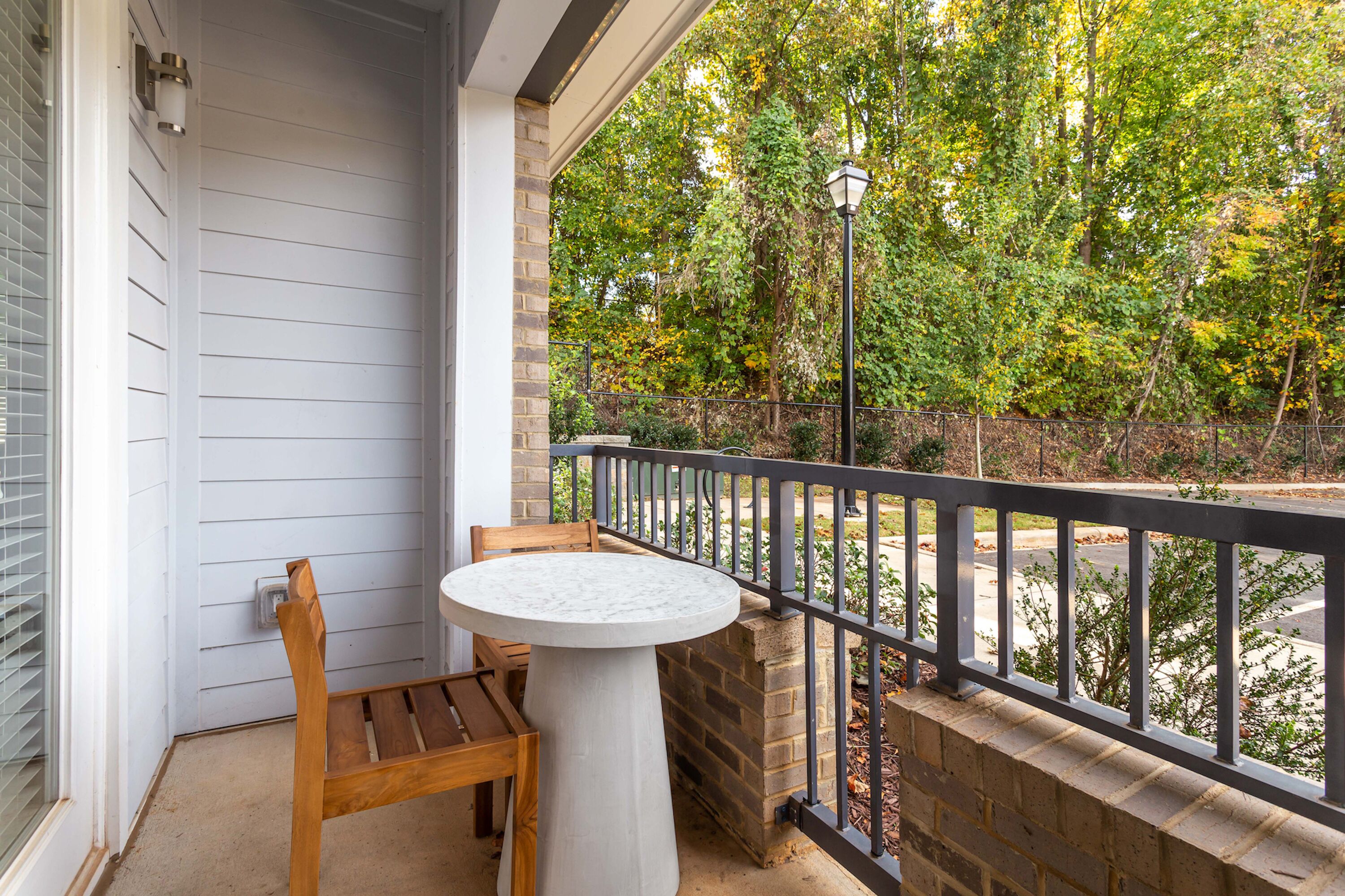Un balcon avec un banc en bois et une petite table, donnant sur un jardin avec des arbres et une clôture.