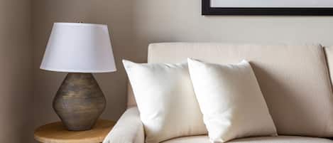 Een beige bank met twee kussens, een houten bijzettafeltje en een lamp met een witte kap.