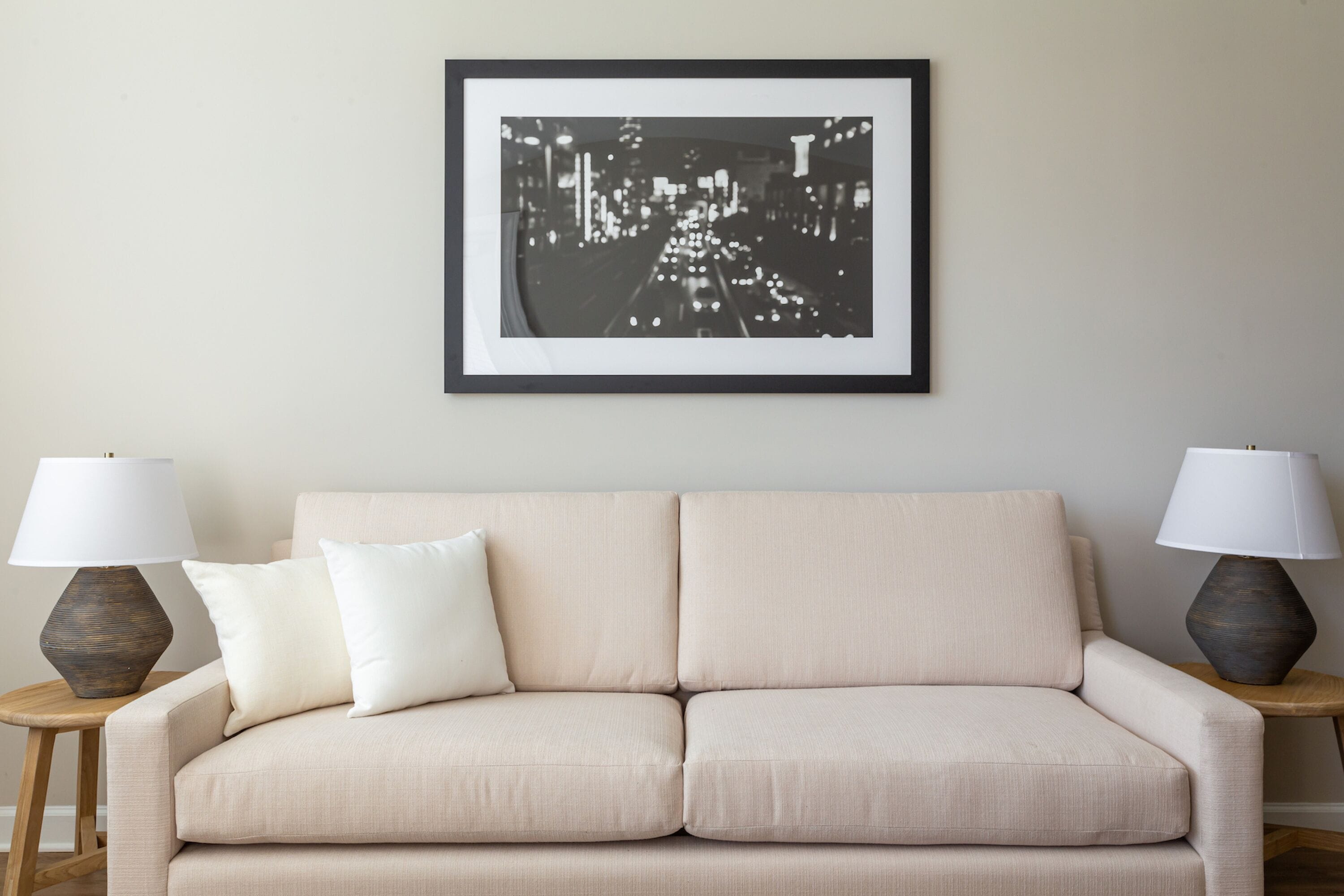 A beige sofa with two lamps on either side, a framed black and white cityscape picture on the wall, and a small wooden side table.