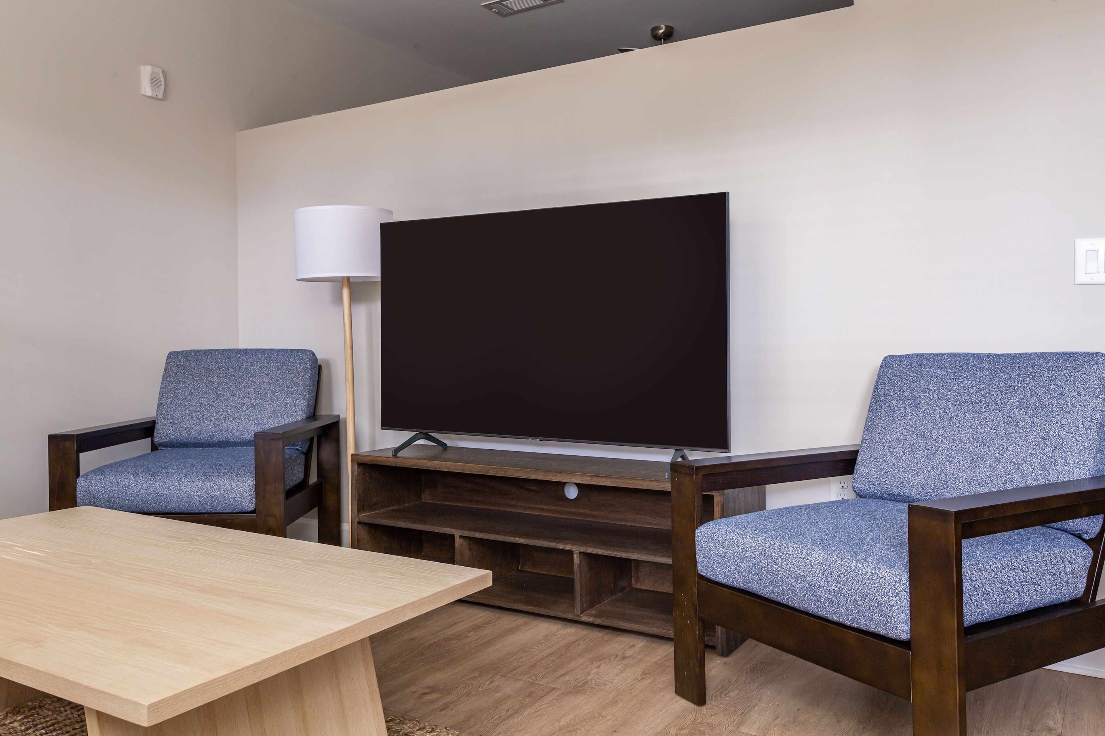 A room with a wooden table, two blue upholstered chairs, a large flat-screen TV, and a lamp.