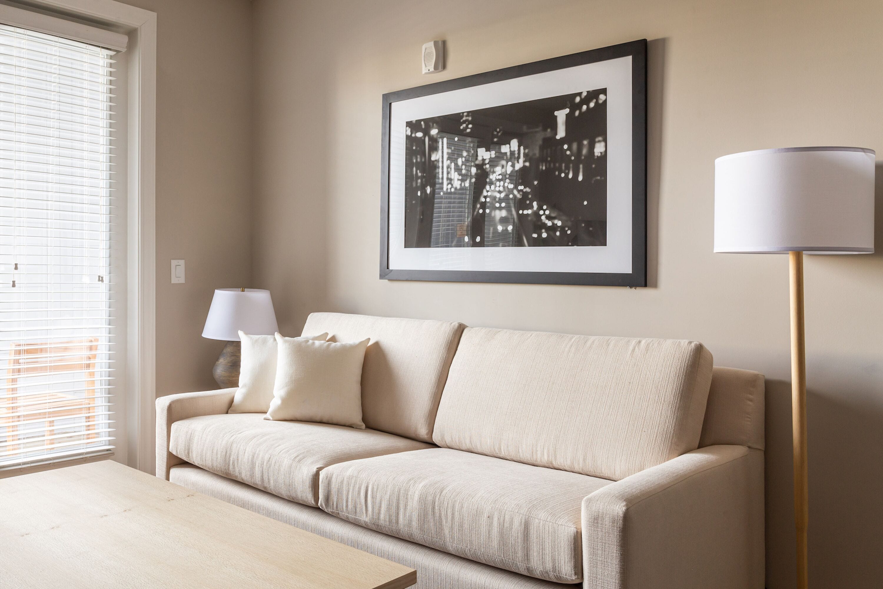 A living room with a beige sofa, a framed picture on the wall, a floor lamp, and a table.