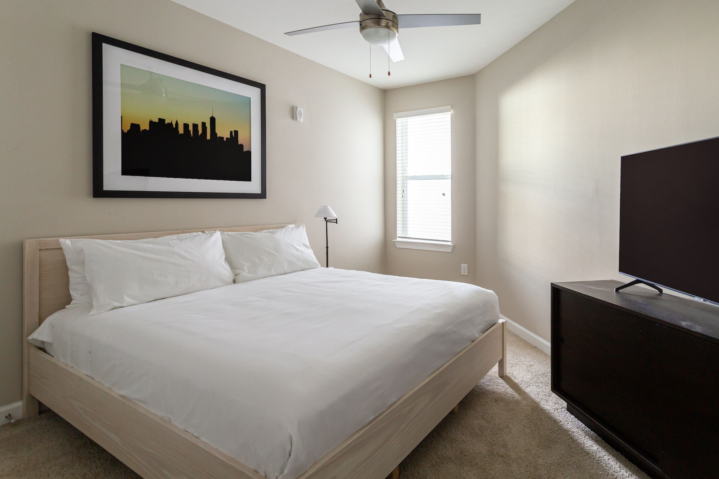 A bedroom with a bed, a ceiling fan, a framed cityscape picture, a window, and a TV on a stand.