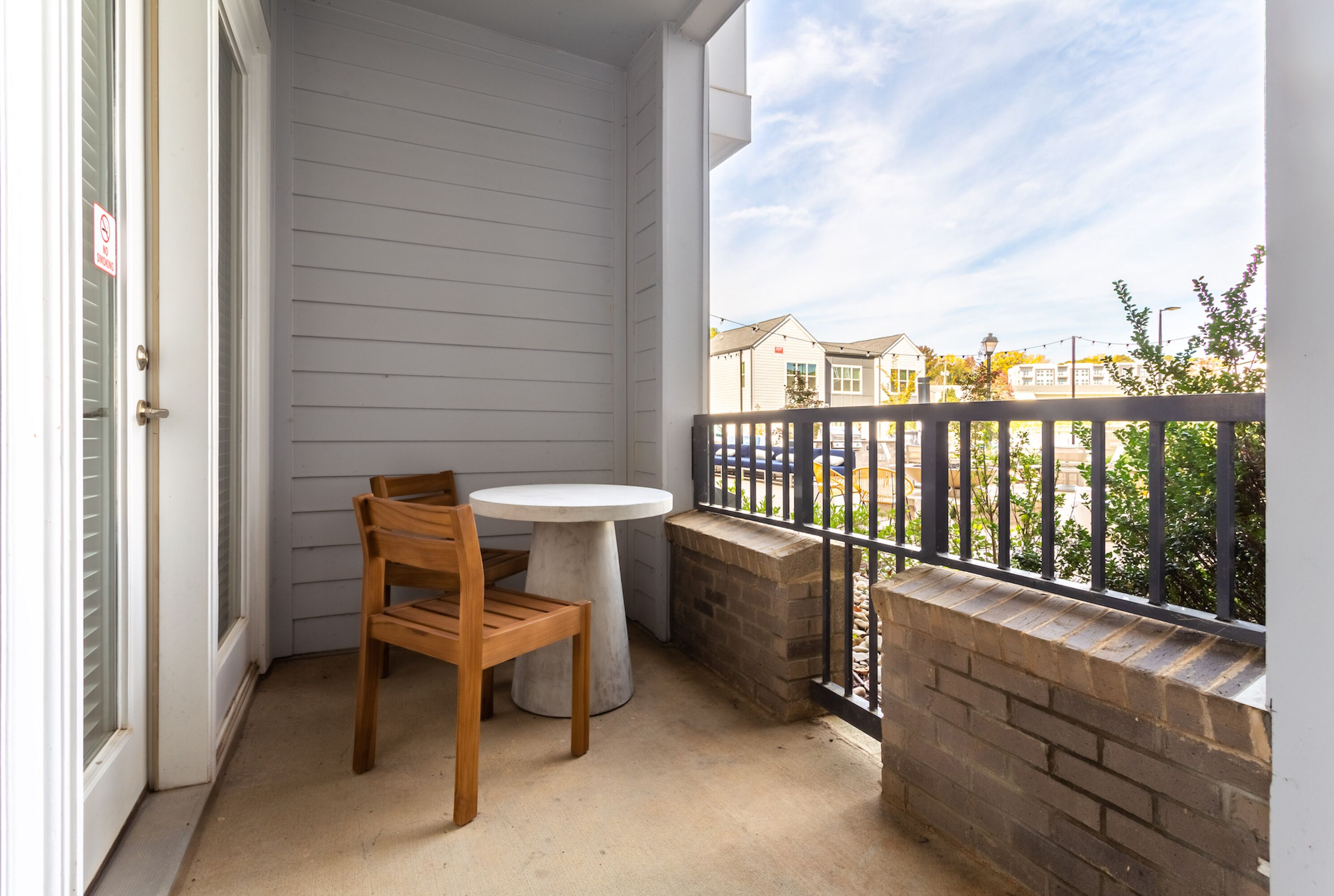 A balcony with a wooden chair and a small table, a brick wall, and a view of neighboring houses.