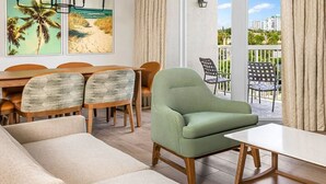 Una sala moderna con un sillón verde, sofá beige, mesa de comedor de madera y sillas. Hay dos ventanas grandes con cortinas, y un balcón con mobiliario exterior.