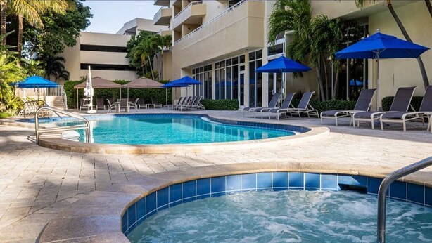 Zona de piscina con sillones de descanso y sombrillas, rodeada por un edificio con balcones.