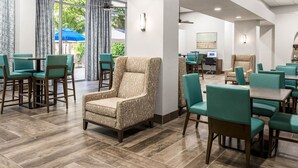 Un vestíbulo de hotel con sillas y mesas color turquesa, un sillón estampado y un ventanal con cortinas.