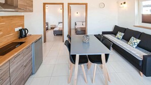 Una cocina moderna con encimera de madera y una máquina de café, una zona de comedor con mesa y sillas, y un salón con sofá y cojines estampados.