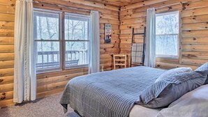 Quarto de cabana de madeira com cama, duas janelas com cortinas, uma placa de boas-vindas e uma escada de madeira.