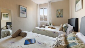 Ein Hotelzimmer mit zwei Betten, einem Fenster mit Vorhängen, einem gerahmten Bild an der Wand und einem Tablett mit einer Flasche und Gläsern auf einem Bett.