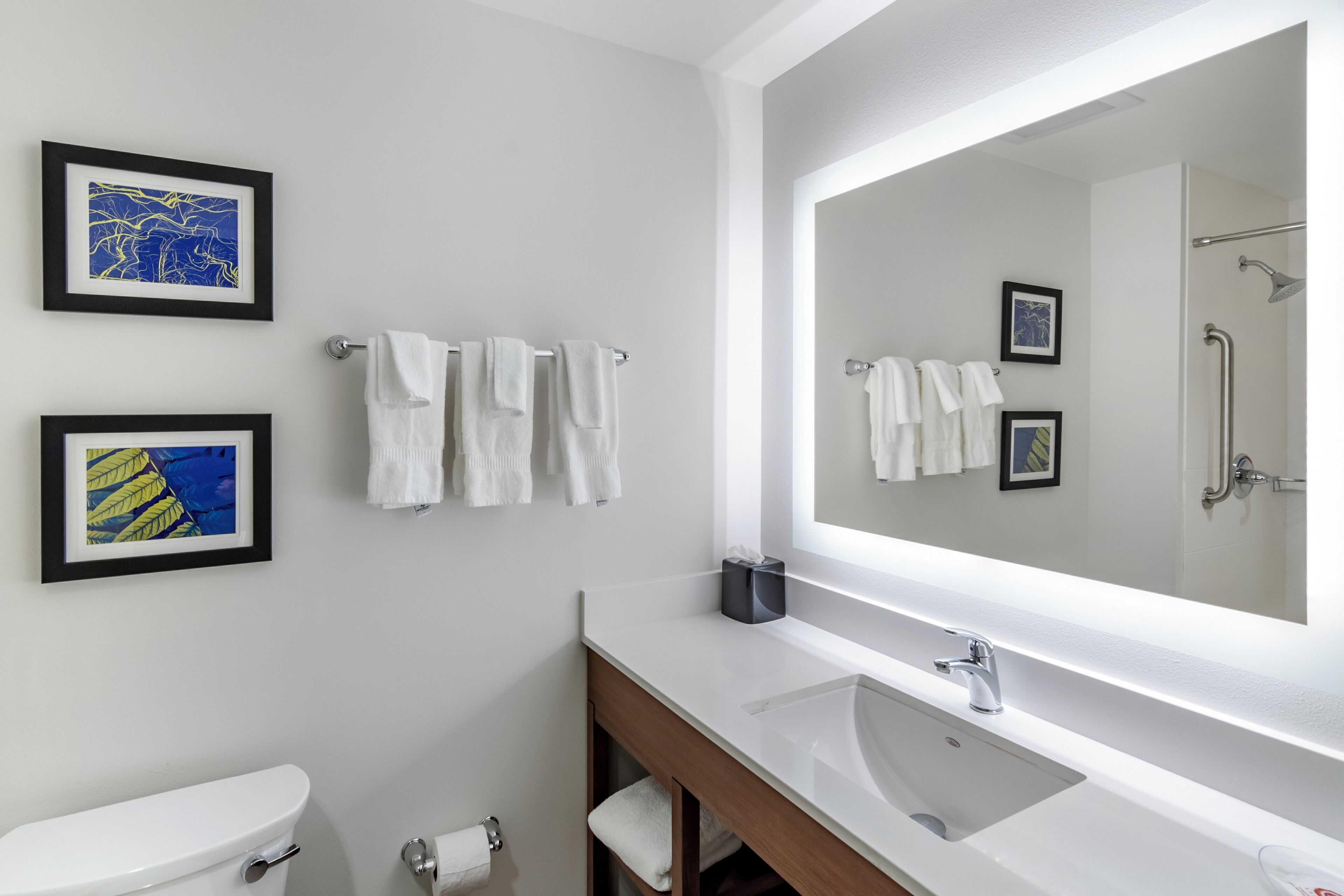 A modern bathroom with a toilet, sink, and large mirror. There are framed pictures on the wall and several white towels.