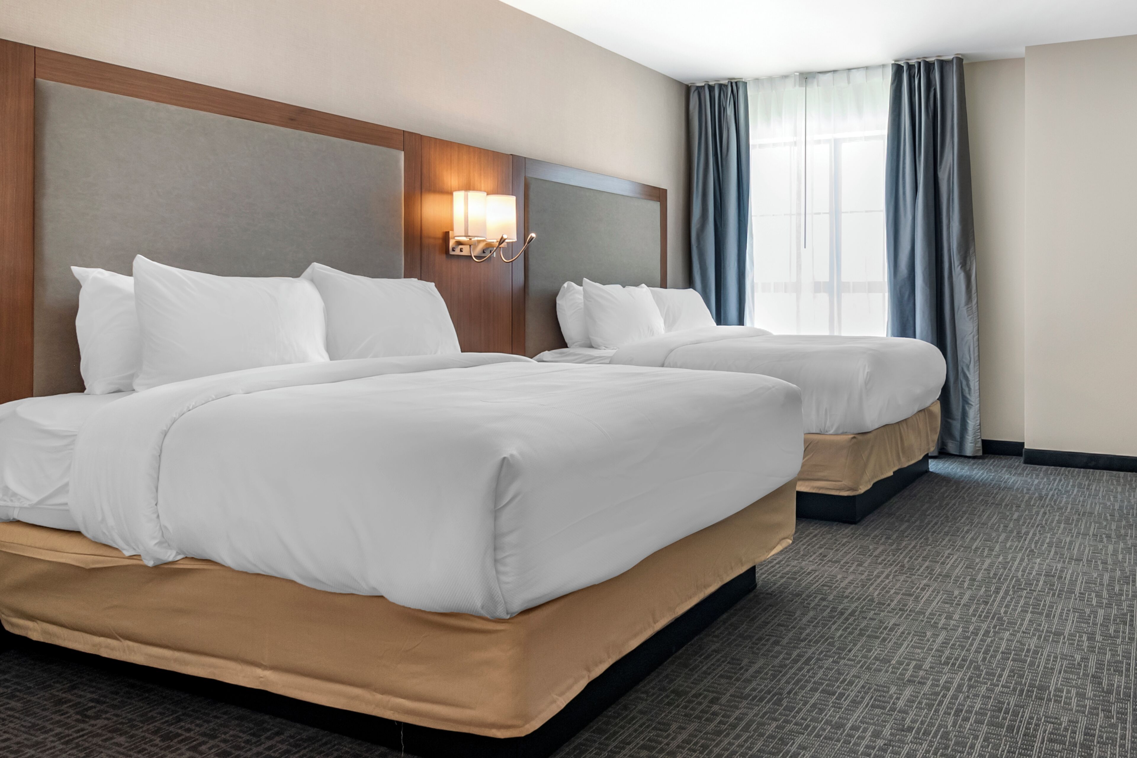 A hotel room with two beds, a headboard with a light fixture, a large window with curtains, and a carpeted floor.