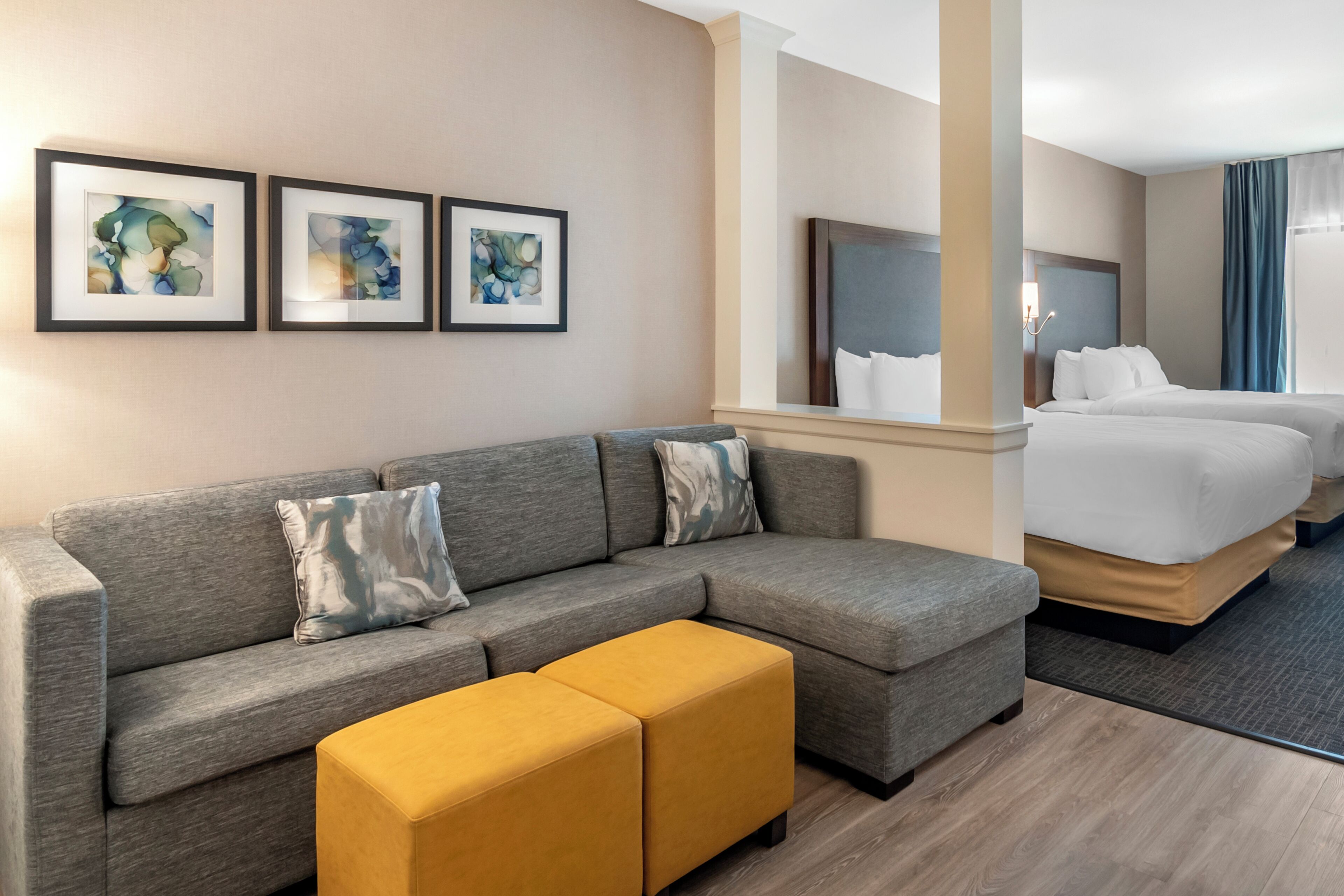 Une chambre d’hôtel avec un canapé gris, un pouf jaune, un lit et trois tableaux abstraits encadrés accrochés au mur.