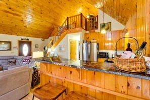 Uma sala de estar espaçosa com teto de madeira, cozinha com armários de madeira, bancada de pedra e escada que leva a um andar superior.
